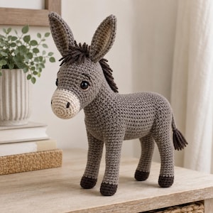 Pode incluir: Um burro de peluche feito em croché com corpo cinzento, focinho bege claro, cascos castanho-escuros e crina castanho-escura. O burro tem orelhas grandes e eretas, olhos de plástico preto e uma cauda pequena com um tufo castanho-escuro.