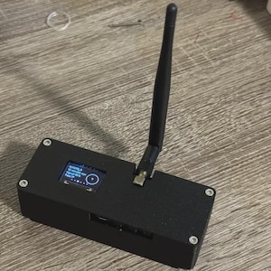 May include: A rectangular black electronic device featuring a small digital display screen showing text and a circular icon, with a single black antenna extending vertically from the top surface, secured by four silver screws at the corners.