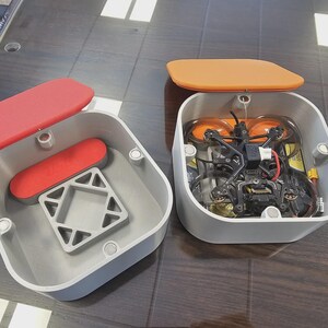 Peut inclure: Deux boîtes de rangement carrées grises aux coins arrondis. L'une possède un couvercle et un insert rouges, tandis que l'autre a un couvercle orange et contient un drone FPV noir avec des hélices orange à l'intérieur de la base.