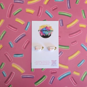 May include: White and gold teacup earrings with pink bows, presented on a white card with the "Charming Clay" logo. The background is a pink surface with colourful, curved shapes.