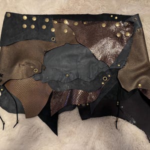 Könnte beinhalten: Ein Rock aus überlappenden Flicken aus schwarzem Wildleder und bronzefarbenem Material mit Schlangenlederstruktur. Das Kleidungsstück ist mit messingfarbenen Metallnieten, Ösen und dünnen schwarzen Lederbändern an den unteren Rändern versehen.
