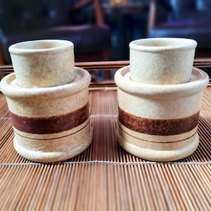 May include: Two cylindrical ceramic cups with a speckled beige finish. Each cup features a wide, dark brown horizontal band around the center, accented by a thin, dark brown line below it. The cups have a thick, rounded rim and a sturdy base.