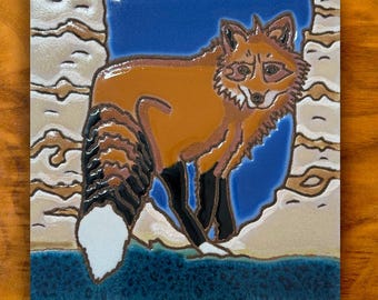 May include: A square ceramic tile featuring a raised relief illustration of a red fox with a bushy tail, black legs, and a white tail tip. The fox is set against a blue background framed by textured, light beige, wavy horizontal bands.