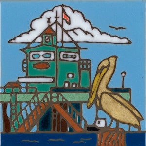 May include: A colorful ceramic tile depicting a green pier building with a red flag, a white cloud, and a yellow pelican standing on a wooden post. The pier is over blue water.