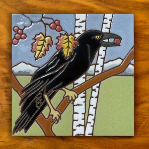 May include: A square ceramic tile featuring a black raven perched on a brown branch. The bird holds a small red berry in its beak. The background displays white birch tree trunks, yellow and red autumn leaves, and distant mountains.