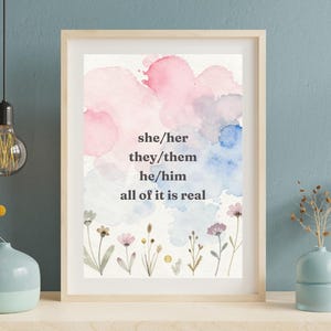 May include: A framed wall art print featuring watercolor splashes in pink, blue, and white. The text reads: she/her, they/them, he/him, all of it is real. Below the text are delicate, hand-painted illustrations of various small wildflowers.