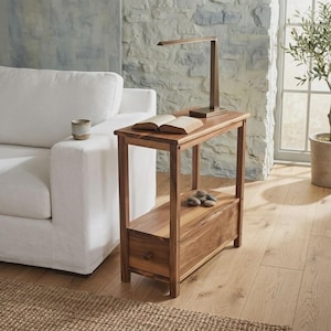 May include: A rectangular wooden side table with a natural grain finish, featuring a flat top surface, an open middle shelf, and a bottom storage drawer with a small round knob. A slim, modern metal desk lamp and an open book sit on the top surface.