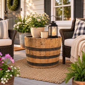 May include: A round wooden barrel table with dark metal bands, featuring a light brown finish. On top of the table sits a white ceramic pot filled with white daisies, a beige speckled ceramic mug, and a black metal lantern with a lit candle.