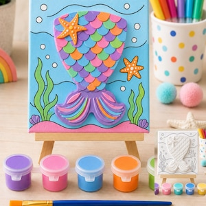 May include: A craft kit featuring a canvas on a wooden easel with a raised mermaid tail design in purple, pink, orange, and green scales. The scene includes painted seaweed, seashells, and two orange starfish. Includes paint pots and a brush.