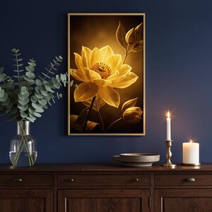 Op de afbeelding: Een rechthoekige wandkunstprint met een gloeiende gele bloem met gelaagde bloemblaadjes en twee gesloten knoppen, ingelijst in goud. Het kunstwerk hangt aan een donkerblauwe muur boven een houten dressoir met een glazen vaas met eucalyptus.