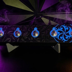May include: A black rectangular guitar effects pedal featuring a geometric triangular pattern across its surface. It includes five metallic footswitches with blue LED rings and a blue circular swirl graphic on the right side of the unit.