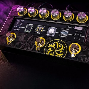 May include: A black guitar effects pedal with a geometric textured surface and yellow accents. It features six silver knobs, three metallic footswitches, and a central digital display showing signal chain icons and the text ANAGRAM on the side.