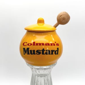 May include: A bright yellow ceramic mustard pot with a matching lid and a wooden spoon. The front of the pot features the text Colman's Mustard in red and black lettering. The pot sits atop a clear, ribbed glass base, creating a distinct contrast.