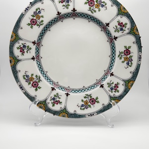 May include: A round ceramic plate featuring a white centre and a wide border decorated with repeating arched panels. Each panel contains small floral clusters in pink, yellow, and blue, framed by a dark teal and black patterned edge trim.