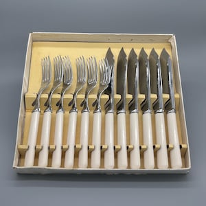 Könnte beinhalten: Ein Set aus zwölf Besteckteilen aus Edelstahl, angeordnet in einem rechteckigen Karton. Das Set enthält sechs Gabeln und sechs Messer, jeweils mit einem polierten silberfarbenen Metallkopf und einem glatten, cremeweißen Griff.