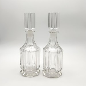 Peut inclure: Deux carafes en verre transparent avec des détails nervurés verticaux sur le corps et des bouchons rectangulaires assortis. Chaque carafe présente une base hexagonale et un col fin, offrant un design transparent aux lignes épurées.