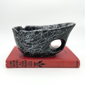 May include: A black ceramic bowl with a white splattered paint pattern and a circular side cutout, resting on a red book titled A TRIP AROUND THE WORLD in a FLYING MACHINE by JULES VERNE. The bowl features a notched rim and a glossy finish.