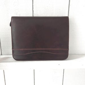 May include: A rectangular dark brown leather portfolio case with a smooth finish and a horizontal stitched seam detail across the lower front section. The case features a flap closure that extends across the top and rounded corners at the base.