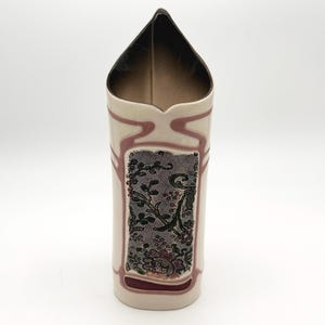 Puede incluir: Un jarrón de cerámica alto de color crema con un borde triangular puntiagudo. El exterior presenta un panel rectangular central decorado con un patrón floral negro similar al encaje, enmarcado por líneas en relieve de color rosa empolvado.