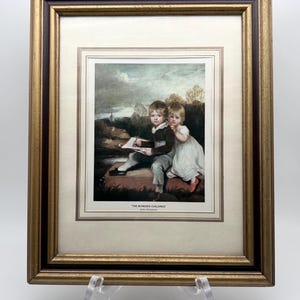 May include: A framed print titled THE BOWDEN CHILDREN by John Hoppner, featuring two children sitting outdoors. One child wears a dark jacket and light trousers, holding a book, while the other wears a white dress. The frame is gold and brown.