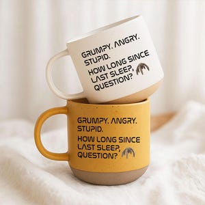 Könnte beinhalten: Zwei vertikal gestapelte Keramikbecher, einer in Creme und einer in Senfgelb, beide mit gesprenkelter Textur und dem schwarzen Aufdruck: GRUMPY. ANGRY. STUPID. HOW LONG SINCE LAST SLEEP, QUESTION? sowie einer kleinen Grafik einer Kreatur.