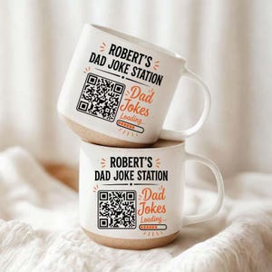 Könnte beinhalten: Zwei cremefarbene Keramiktassen mit gesprenkelter Oberfläche und beigem Boden. Jede Tasse zeigt den schwarzen Schriftzug "ROBERT'S DAD JOKE STATION", einen schwarzen QR-Code und den orangefarbenen Text "Dad Jokes Loading..." mit Ladebalken.