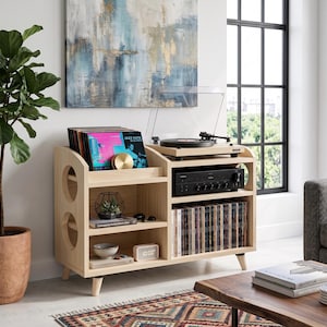 Puede incluir: Una consola multimedia de madera clara con una superficie superior para un tocadiscos, una ranura para discos de vinilo y varios estantes abiertos. La unidad incluye un receptor electrónico negro, un reloj digital, libros y objetos decorativos.