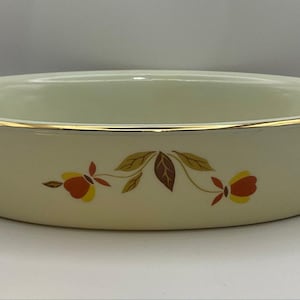 May include: An oval-shaped cream-colored ceramic baking dish featuring a gold-trimmed rim and a decorative floral pattern in shades of orange, yellow, and brown. The dish has a smooth, glossy finish and a shallow, wide-rimmed design.