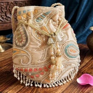 May include: A beige drawstring pouch bag featuring intricate embroidery in shades of orange, teal, and gold. The bag is adorned with small white beads, gold metallic thread, and a decorative tassel with hanging pearl-like beads at the center.