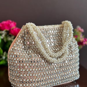 Könnte beinhalten: Eine pfirsichfarbene Handtasche mit einer aufwendigen Außenseite, die mit kleinen, reflektierenden, rautenförmigen Spiegeln und weißen Kunstperlen besetzt ist. Die Tasche hat zwei Griffe, die aus mehreren Strängen weißer Kunstperlen bestehen.