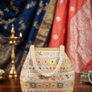 Peut inclure: Un sac à main couleur crème doté d'une anse en perles et de broderies complexes dans des tons pastel de rose, jaune et bleu. Le sac est orné de petits miroirs circulaires disposés en rangées horizontales et de cadres carrés.