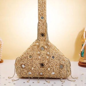 May include: A beige triangular handbag featuring intricate embroidery, embedded square mirrors, and a single top handle. The bag is adorned with dangling beaded chains and small white pearls, creating a textured surface with a symmetrical pattern.