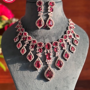 Puede incluir: Un conjunto de joyería compuesto por un collar y pendientes a juego. Las piezas presentan piedras de color rojo intenso, con tallas en forma de pera y ovalada, rodeadas de pequeños cristales transparentes en una montura de metal plateado.