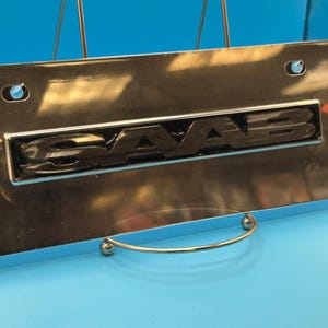 May include: A rectangular chrome license plate frame featuring the word SAAB in raised, glossy black lettering centered on the plate. The metal surface is highly reflective, and the frame includes two circular mounting holes at the top corners.