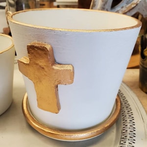 May include: White ceramic planter with a gold cross design and a gold rim. The planter is sitting on a white surface.