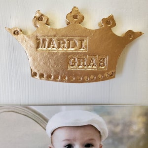 May include: A gold-colored crown with the words "MARDI GRAS" embossed on it. The crown is mounted on a white surface. Below the crown is a photograph of a baby wearing a white hat.