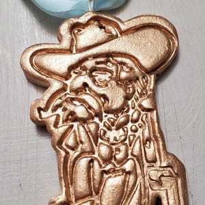 May include: A bronze-colored ornament in the shape of a cowboy with a hat, beard, and rifle. The ornament is suspended by a light blue ribbon. The cowboy figure has detailed features and is likely a decorative item.