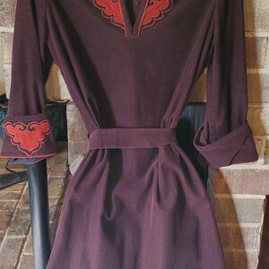 May include: A burgundy dress with long sleeves and a mandarin collar. The dress has red embroidered details on the collar and cuffs. The dress has a belt and is made of a textured fabric.