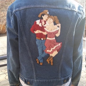 May include: A dark blue denim jacket with a colorful embroidered design of a couple dancing on the back. The embroidery features a man in a red and white striped shirt and a woman in a pink dress.