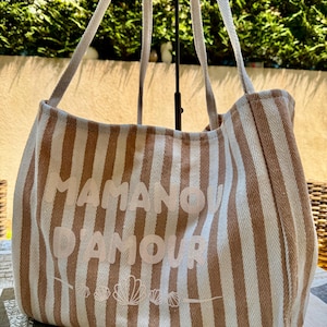 Peut inclure: Un sac fourre-tout avec un motif à rayures verticales beige et blanc et une texture à chevrons. L'avant présente le texte MAMAN D'AMOUR en lettres claires au-dessus d'une ligne graphique composée de cinq petites illustrations de coquillages.