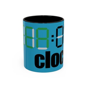 Peut inclure: Un mug en céramique bleu vif avec un intérieur noir. L'extérieur présente un grand graphique d'affichage d'horloge numérique en vert, blanc et noir, avec le mot "clock" imprimé en lettres minuscules noires grasses au bas de la surface.