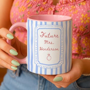 Puede incluir: Una taza de cerámica con asa e interior de color rosa claro, con un patrón de rayas verticales azul claro en el exterior. Un marco rectangular blanco central contiene el texto rosa Future Mrs. Henderson sobre un pequeño icono de anillo.