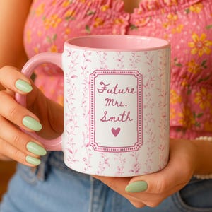 Puede incluir: Una taza de cerámica blanca con interior y asa de color rosa, con un patrón repetitivo de enredaderas florales en rosa claro. El centro muestra un marco rectangular con el texto Future Mrs. Smith en letra cursiva rosa y un pequeño corazón.