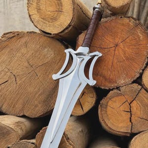May include: A silver-colored metal sword featuring a double-edged blade with a pointed tip and a stylized, curved crossguard. The handle is wrapped in brown material, and the pommel is a simple silver-colored metal cap at the end of the grip.