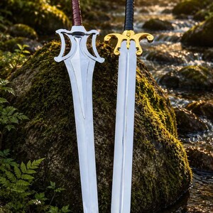 May include: Two metal swords standing upright. The left sword features a silver blade, a brown leather-wrapped handle, and a wide, angular silver crossguard. The right sword has a silver blade, a black handle, and a gold-colored ornate crossguard.
