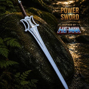 May include: A silver-colored metal sword with a brown wrapped handle and a distinctively shaped crossguard. The blade is long and pointed. Text on the image reads: POWER SWORD INSPIRED BY HE-MAN. The sword is positioned against a large mossy rock.