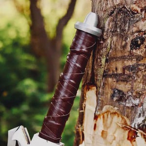May include: A cylindrical handle wrapped in dark brown leather strips with a silver-colored metal pommel at the top. The handle features a spiral pattern created by the overlapping leather wrap, ending in a metallic cap with a rounded top.