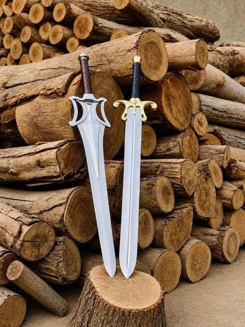 May include: Two metal swords with pointed blades. The left sword features a silver-toned guard and a brown wrapped handle. The right sword has a gold-toned guard with a central emblem and a black wrapped handle. Both blades are double-edged.
