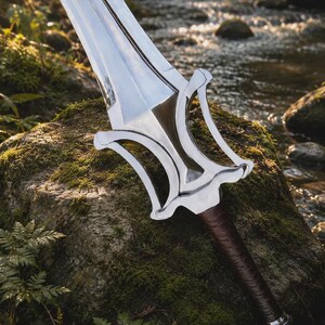 May include: A silver-colored metal sword with a double-edged blade and a complex, open-work crossguard. The handle is wrapped in dark brown leather, and the pommel is a simple silver-colored metal cap. The blade reflects light clearly.