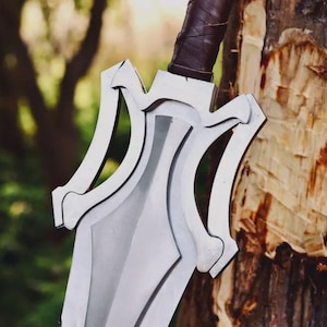 May include: A silver-colored sword with a wide, pointed blade and a complex, multi-layered guard featuring symmetrical cutouts. The handle is wrapped in dark brown leather, providing a textured grip for the hilt of this metallic weapon.